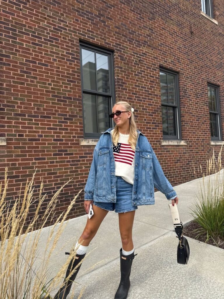 Woman wearing an American flag sweater, denim jacket, shorts, and black rubber boots.