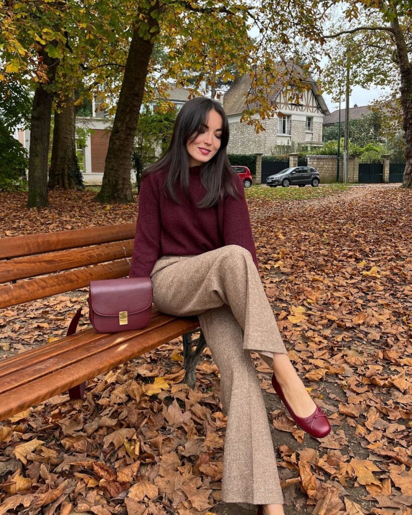 Woman in burgundy sweater and tan trousers sitting on a park bench in autumn