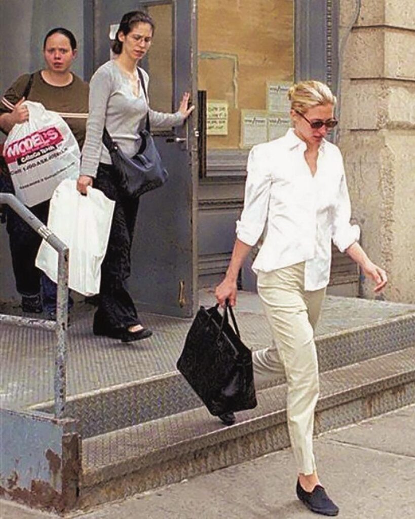 Woman in oversized white shirt and beige pants carrying a large black leather bag.