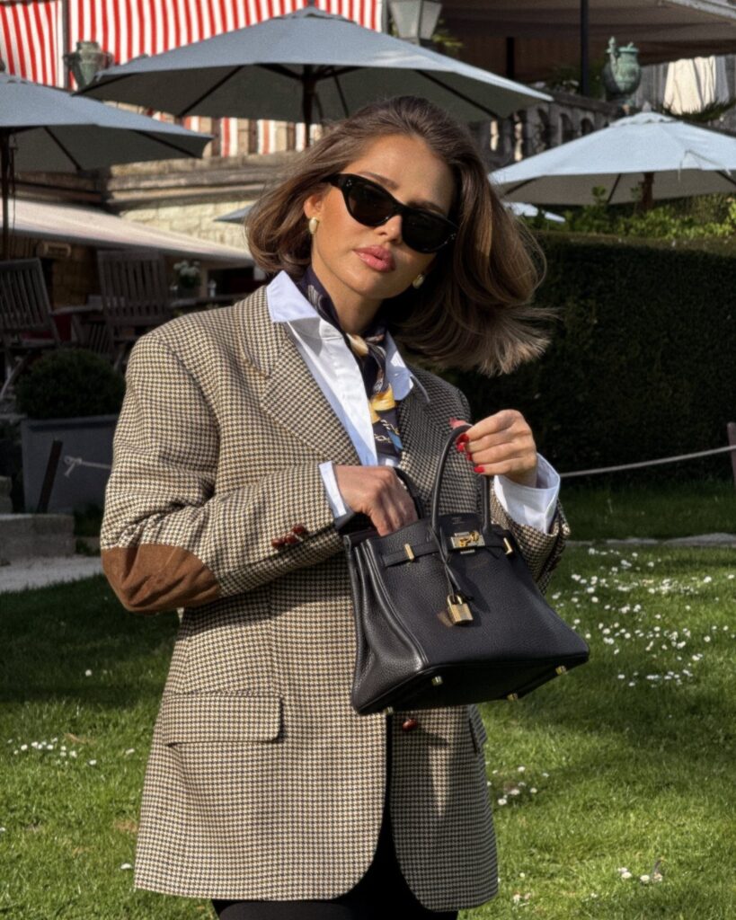 Woman in a checkered blazer and white shirt holding a black leather handbag.
