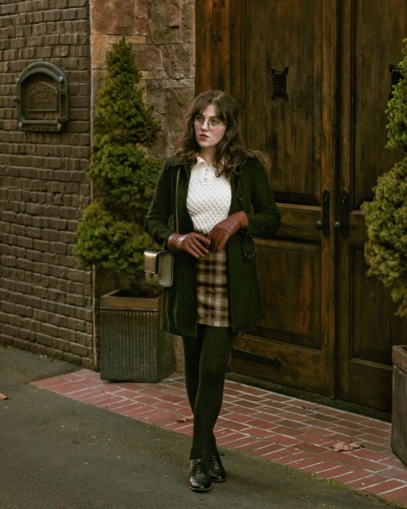 Woman wearing a dark green coat, plaid skirt, and knit sweater in academic style.