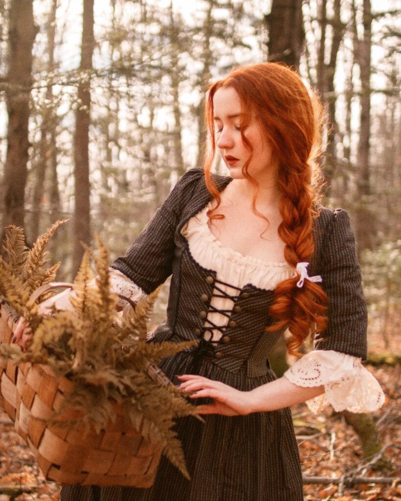 Woman with red hair in a dark pinstriped dress holding ferns in a forest.
