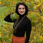 Woman in black turtleneck and rust pleated skirt posing among autumn leaves.