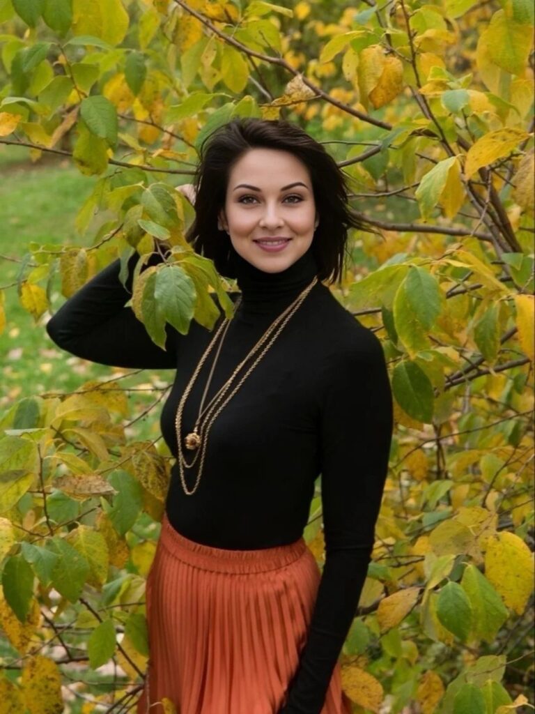 Woman in black turtleneck and rust pleated skirt posing among autumn leaves.