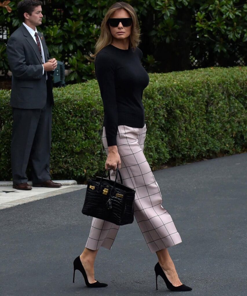 Woman in black top and pink plaid trousers walking outdoors with black handbag