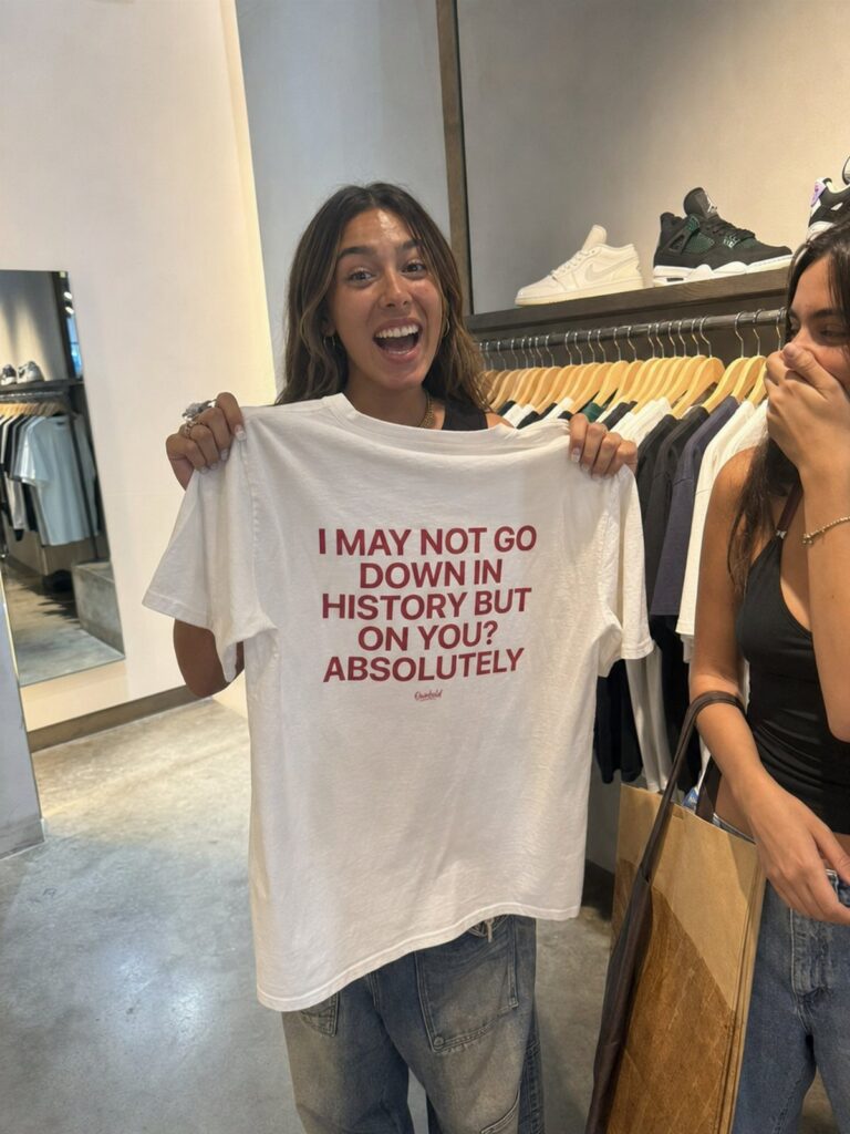 Woman holding a white graphic t-shirt with red text in a clothing store.