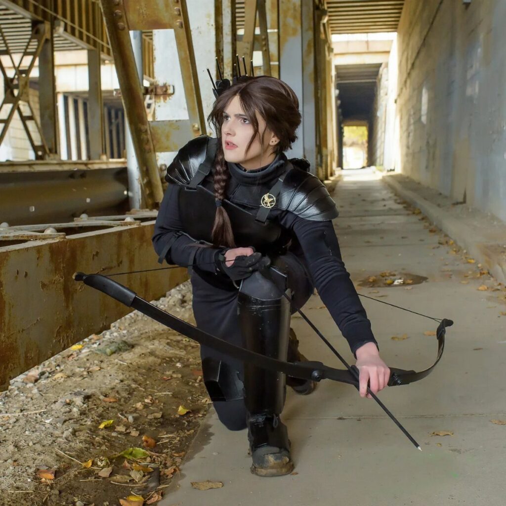 Person in black dystopian huntress costume with bow kneeling in industrial setting.