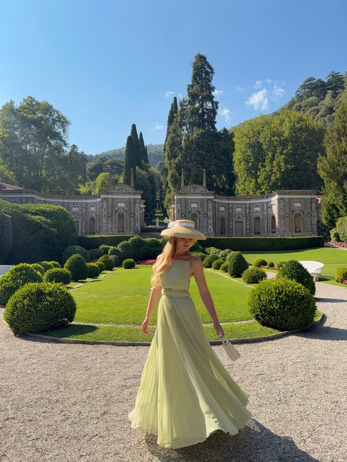 Woman in a long pastel green dress and straw hat in a formal garden.