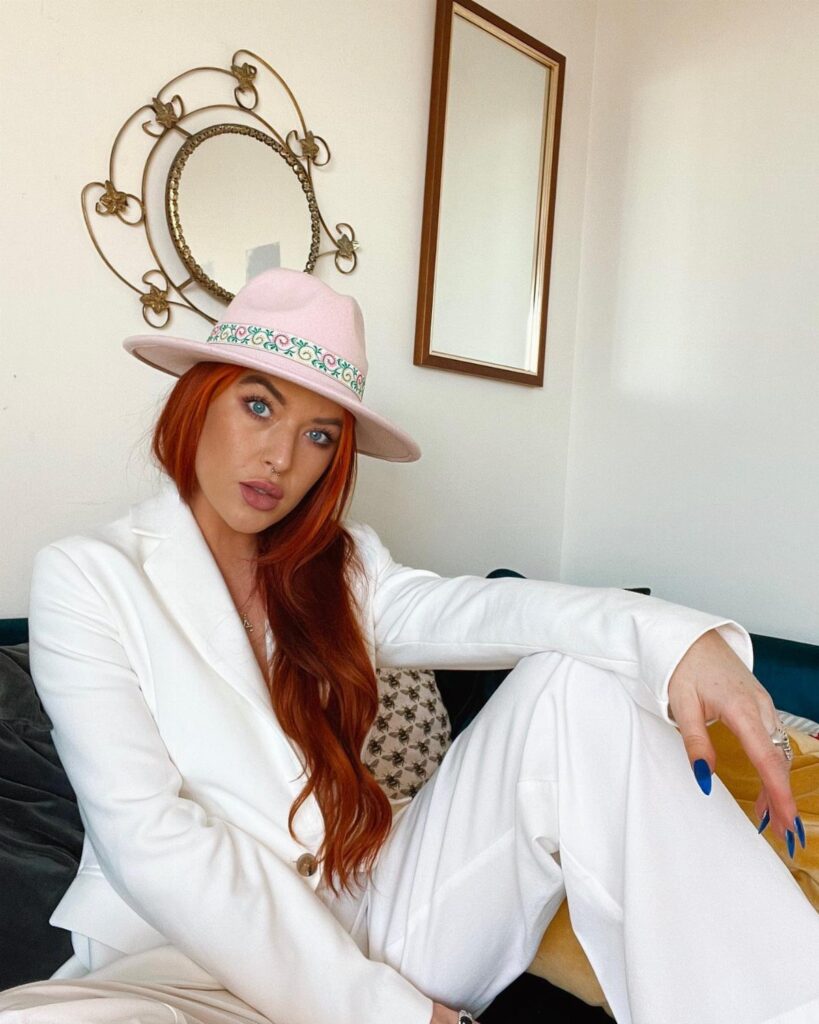 Woman with red hair wearing a pink wide-brimmed hat and white suit.