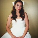 Woman in a white strapless dress and hair bow posing against gold wall.