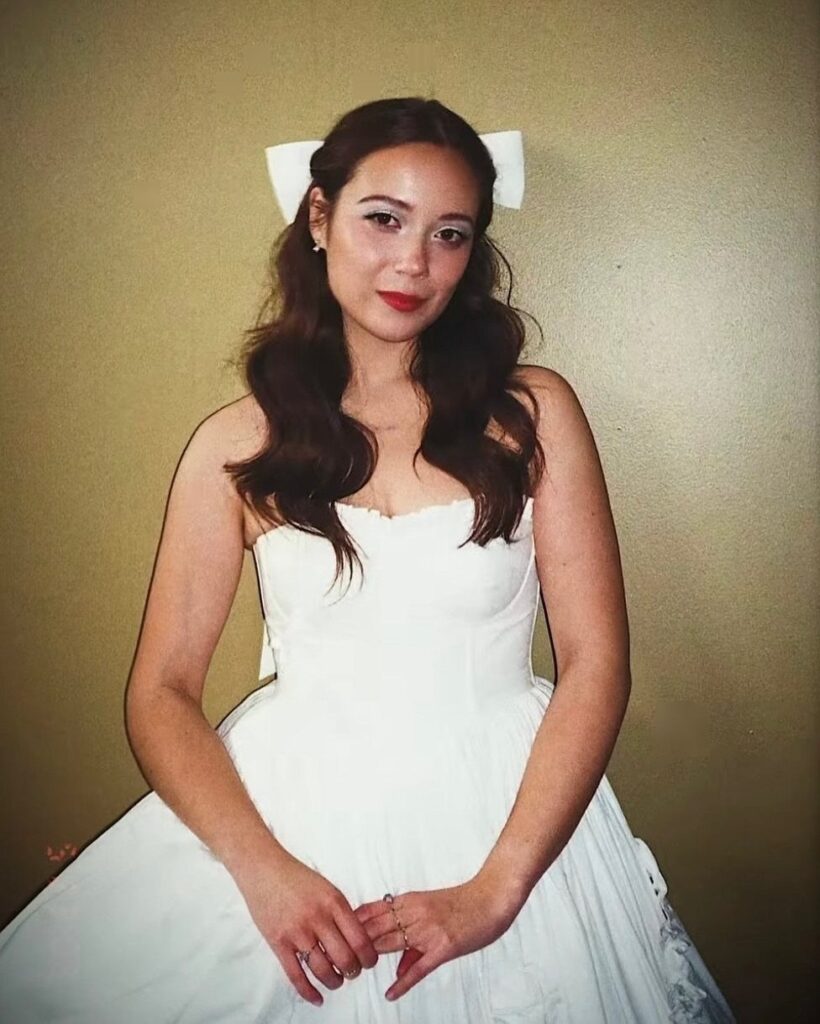 Woman in a white strapless dress and hair bow posing against gold wall.