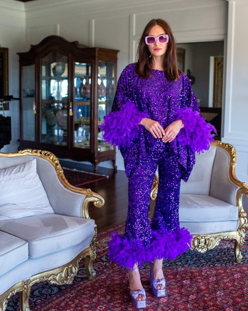 Woman in purple sequined outfit with purple feather trim and pink sunglasses.