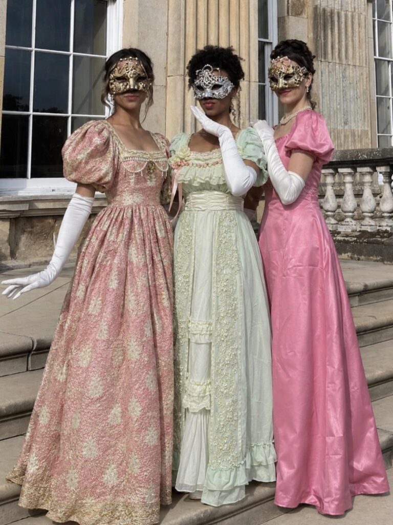Three women in floor-length gowns and ornate masquerade masks posing on stone steps.