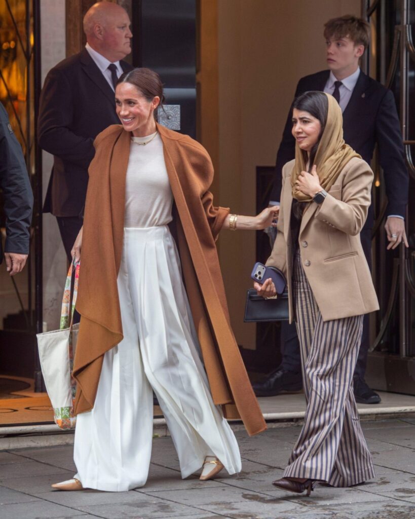 Meghan Markle wearing a white outfit with a long brown coat walking outdoors.