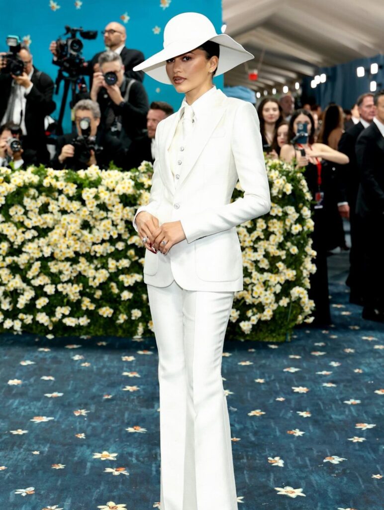 Zendaya in a white tailored suit and wide-brimmed hat on a blue carpet.