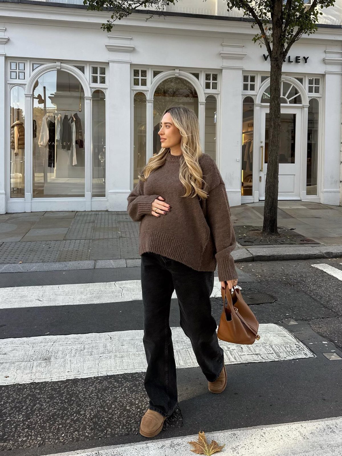 Pregnant woman wearing a brown oversized sweater and black wide-leg trousers on a city street.