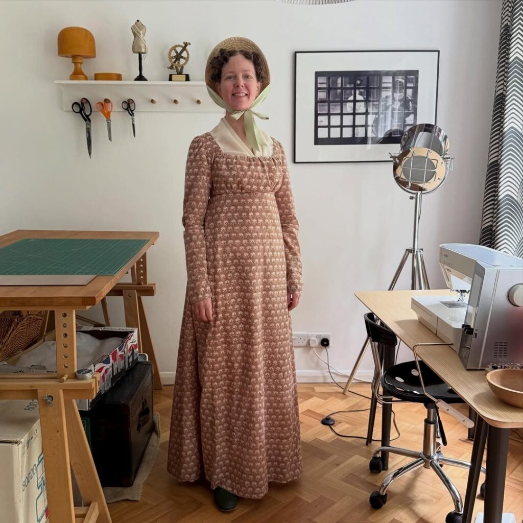 Woman in a brown empire waist Regency dress and straw bonnet in a studio.