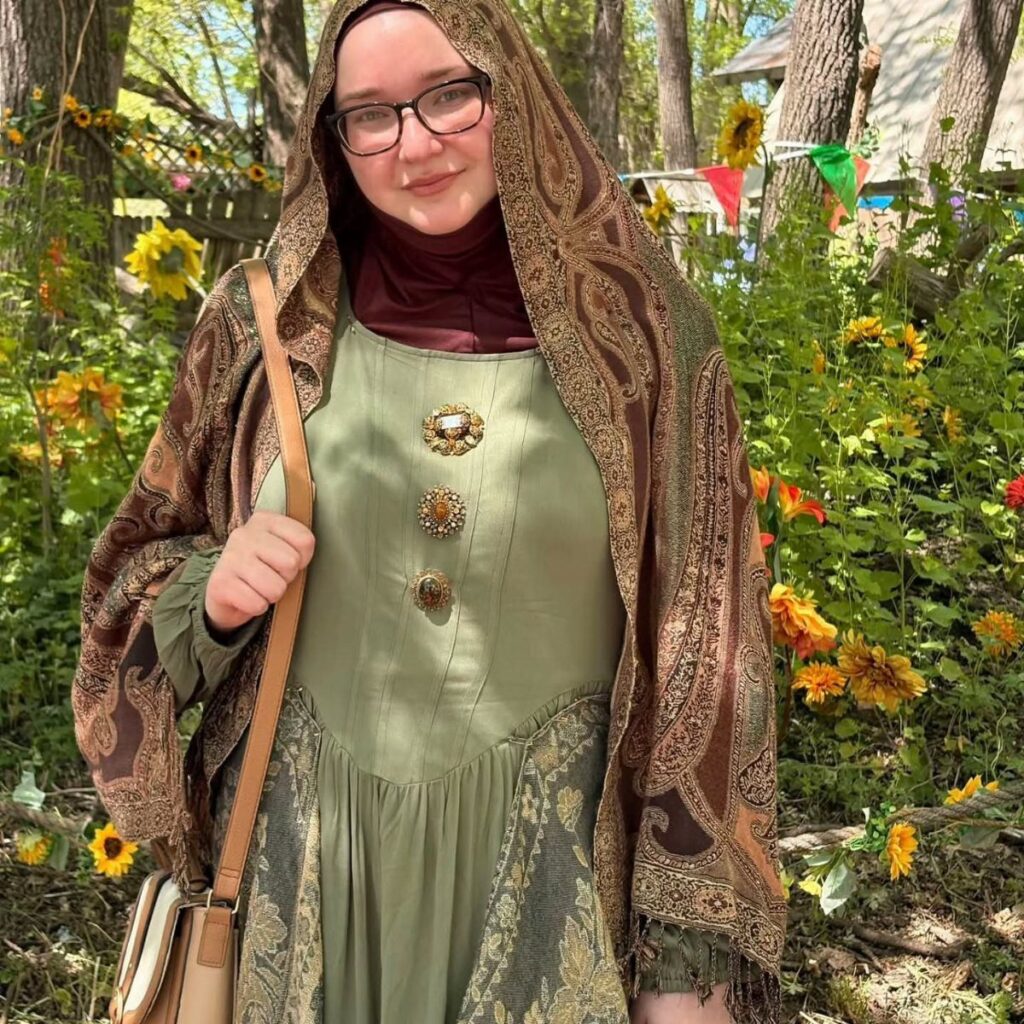 Woman in a green Renaissance dress and patterned brown shawl among yellow sunflowers.