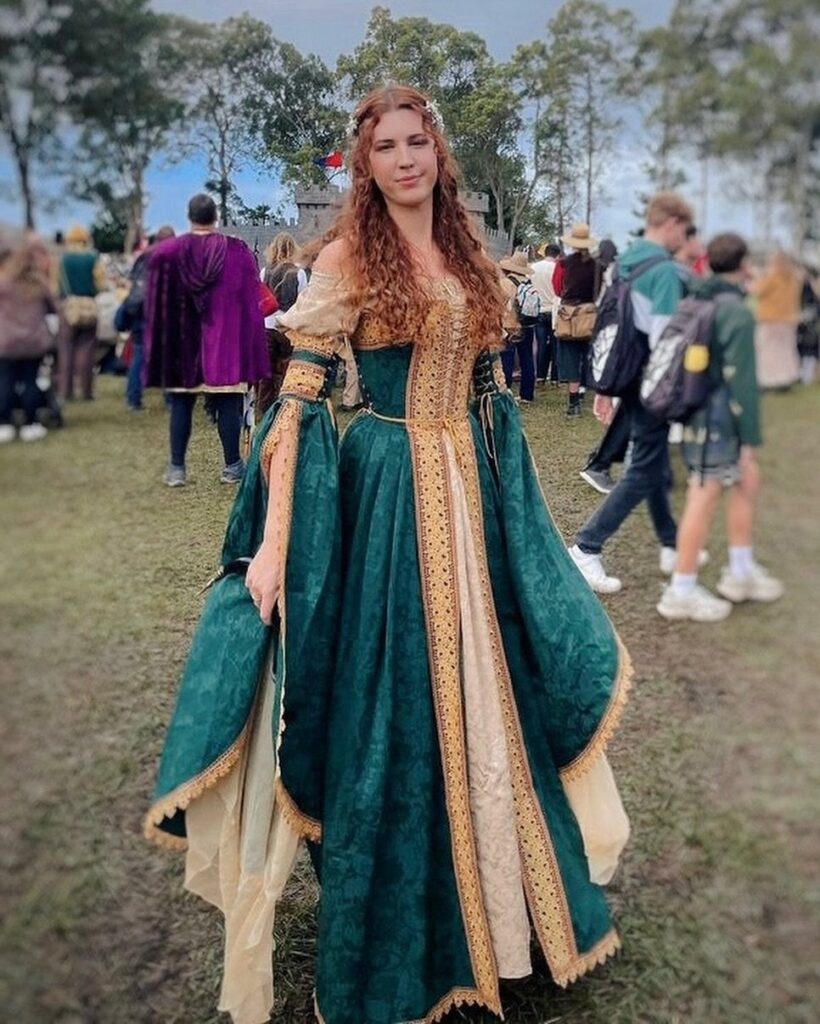 Woman wearing a green and gold Renaissance gown standing in a grassy field.