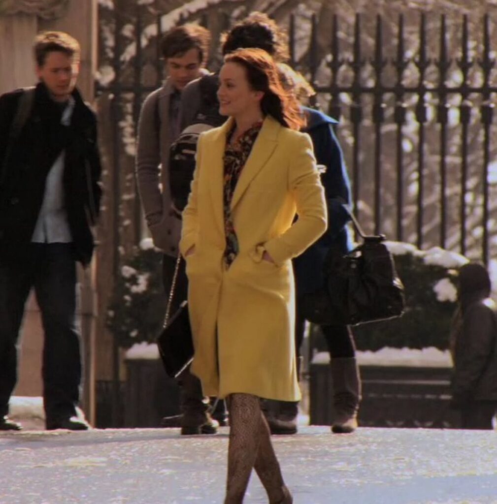 Woman in a bright yellow coat and patterned scarf walking in snowy outdoors.