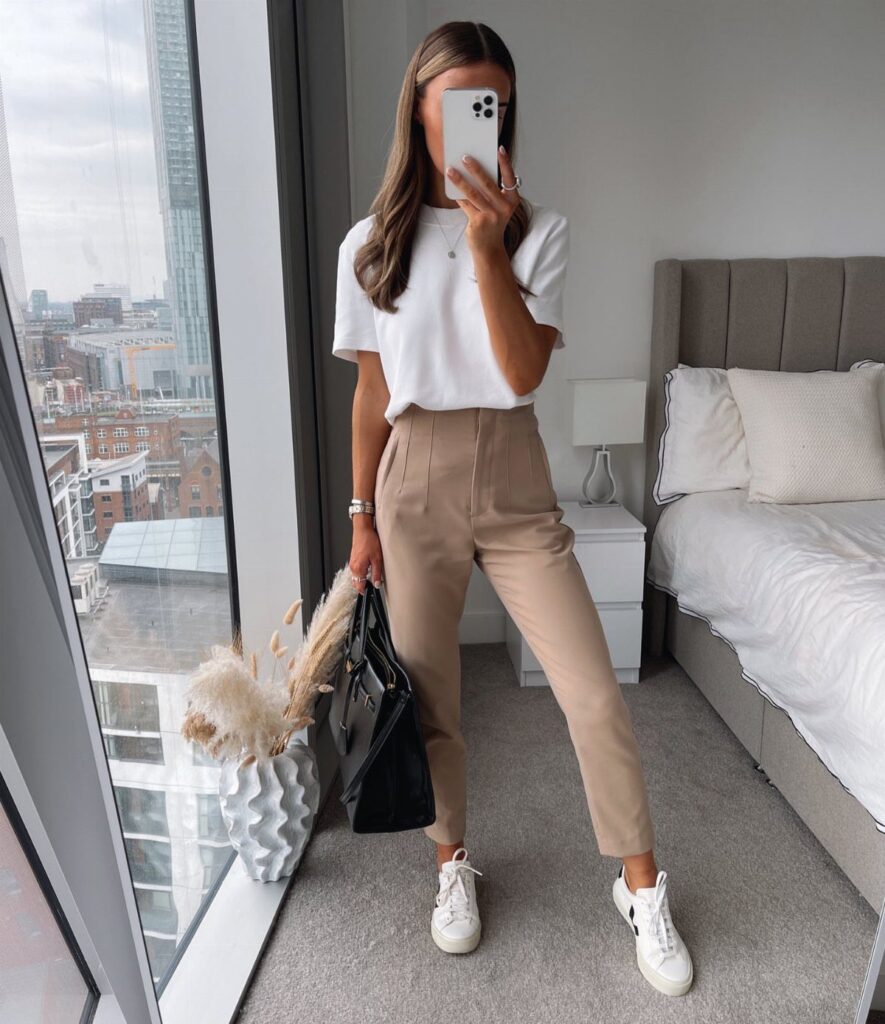 Woman wearing tan tailored trousers, a white t-shirt, and white sneakers in a bedroom.