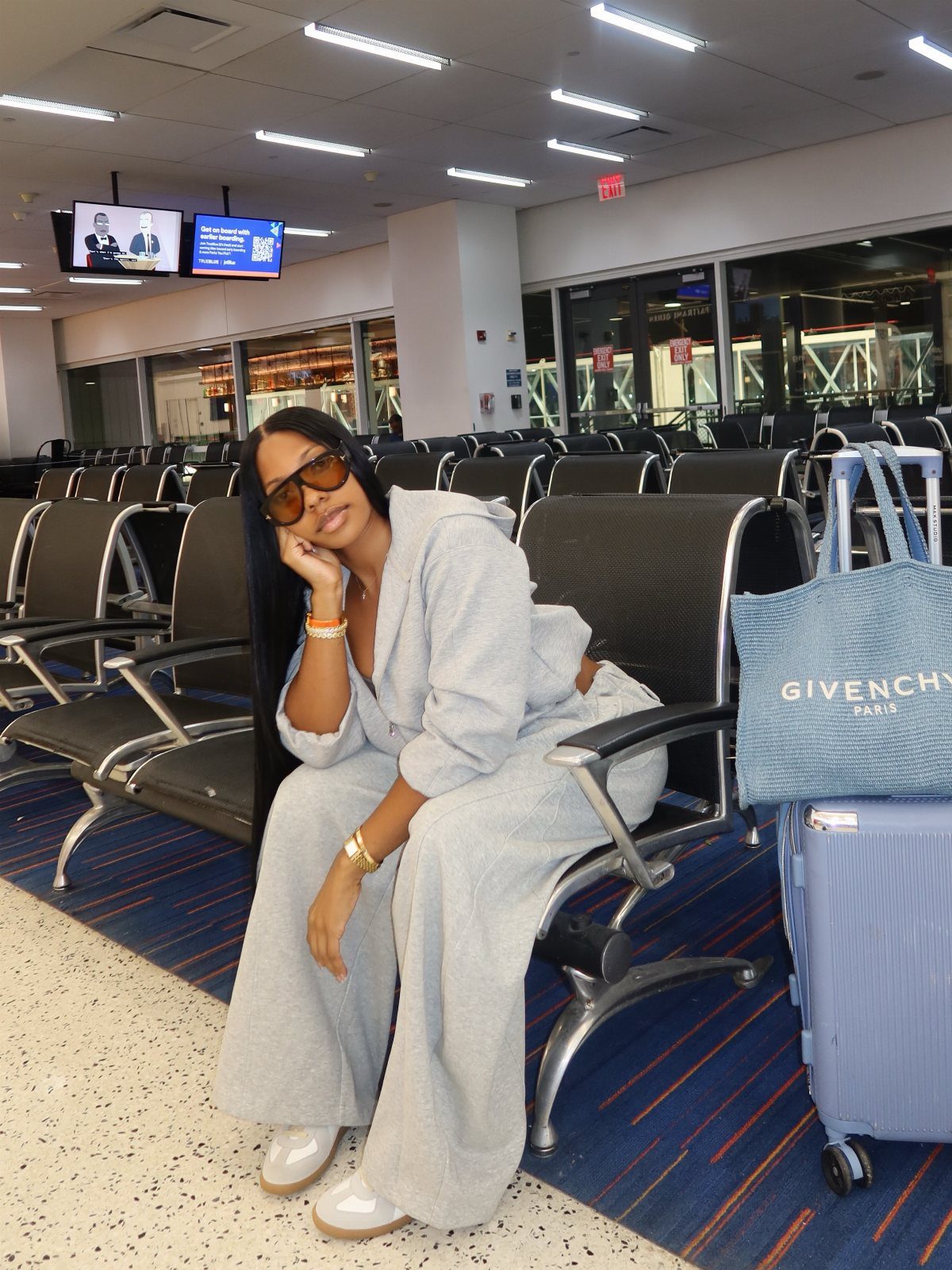 Woman in a grey sweatsuit sitting in an airport terminal with luggage.