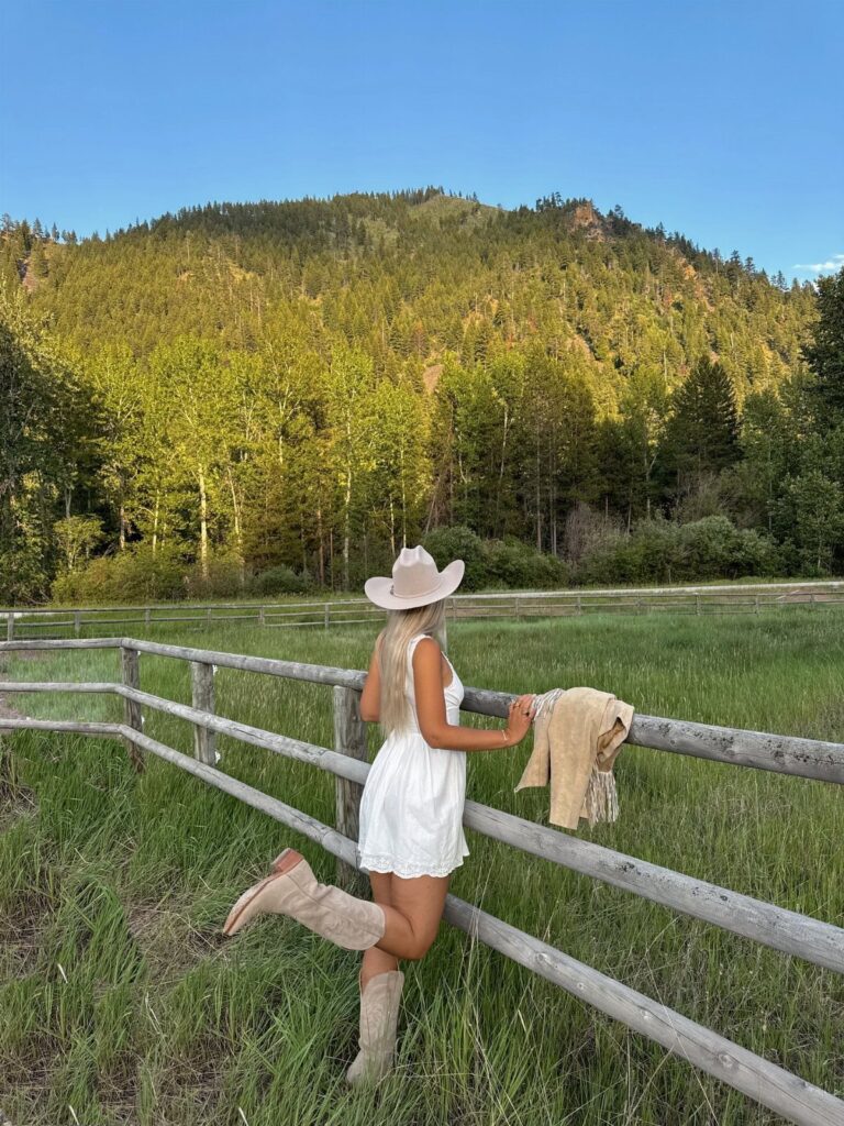 Woman in white dress, cowboy hat, and boots leaning on a wooden fence.